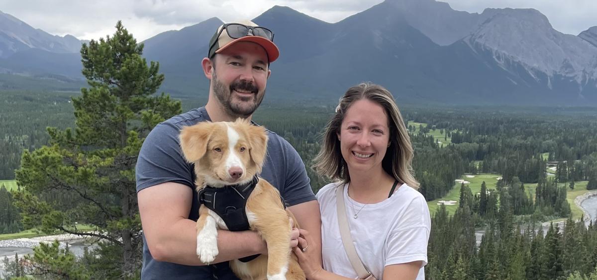Kyle Hager Man holds puppy, woman stands next to him outside with mountains and river in background.