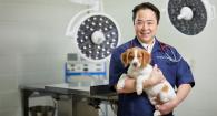Dr. Darin Hisanaga wearing a doctor's smock and stethoscope holds a white and gold puppy in a veterinary hospital exam room.