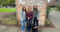 Three women and one black lab dog in front of sign that says Guide Dogs for the Blind