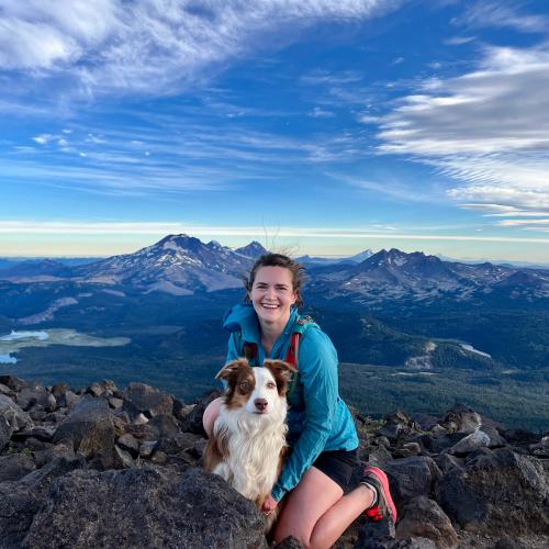 Maggie Mysz and dog on top of a mountain with other mountain peaks in background.